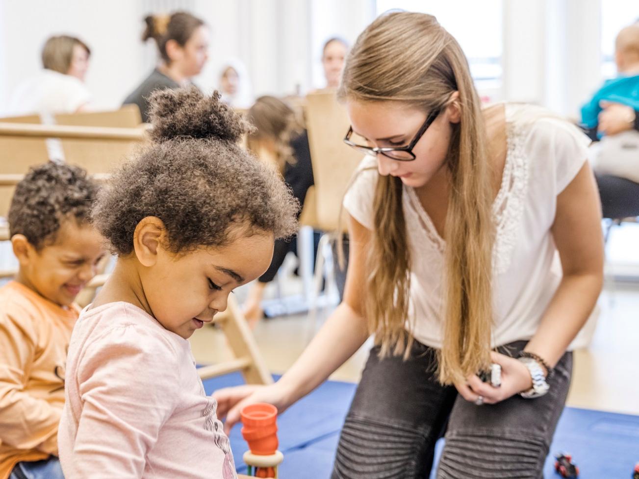 Eltern und KInder im Eltern-Kind-Kurs, im Vordergrund eine Mutter mit Kindern beim Spiel