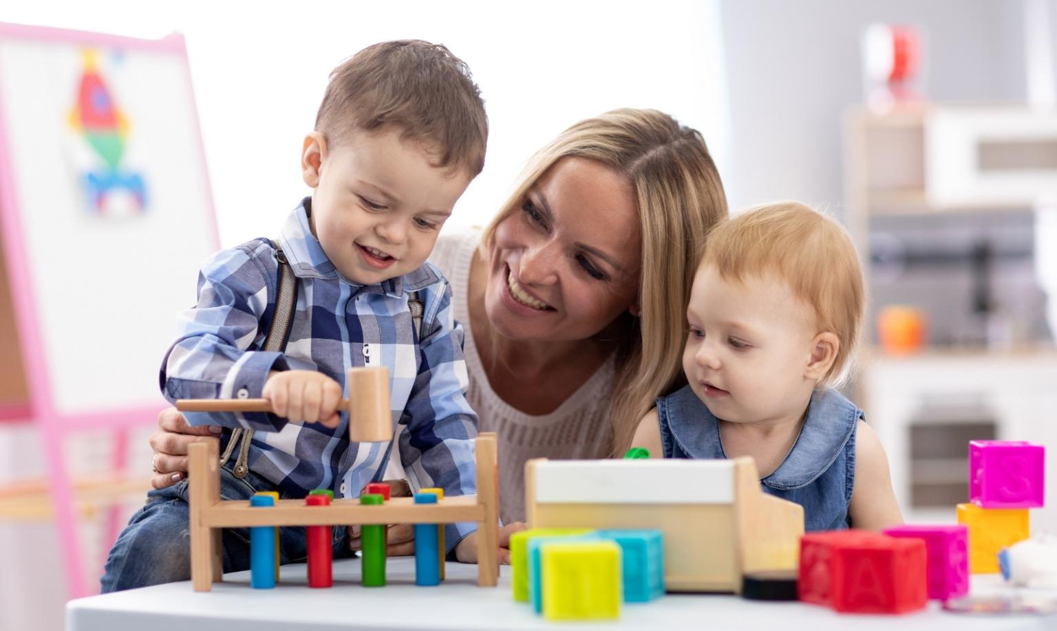 Kinderbetreuerin mit zwei Kleinkindern mit Holzspielzeug am Tisch